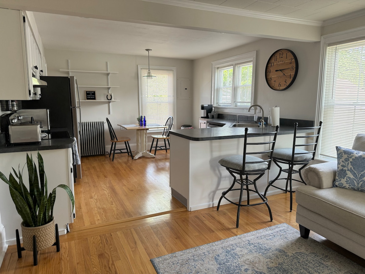 An open-concept kitchen and living area features a dark countertop with bar stools and a dining table set for two. Natural light fills the space through multiple windows, illuminating wood flooring. A cozy couch and decorative plant complete the welcoming atmosphere.