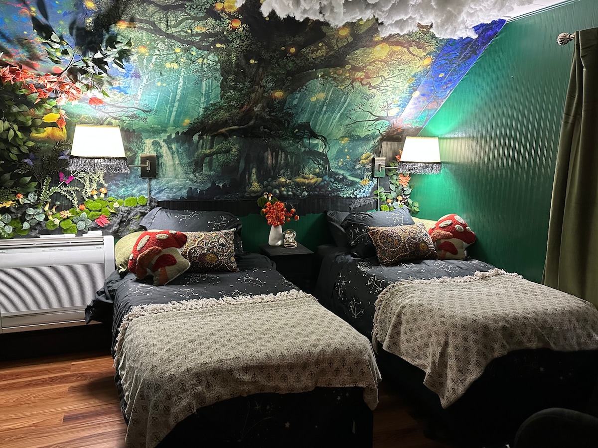 A vibrant room features two twin beds adorned with decorative throw pillows and cozy blankets. A large mural of a mystical tree covers the wall, surrounded by foliage. Soft lamps provide illumination, while a decorative vase adds a touch of color to the nightstand.