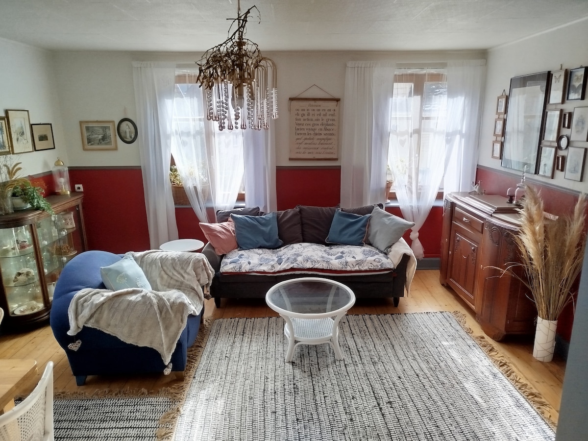 A spacious living area features a mix of vintage furnishings, including a dark sofa adorned with cushions and a cozy throw. A round coffee table sits on a textured rug, while a glass cabinet displays curiosities. Light filters through flowing white curtains, creating a bright atmosphere.