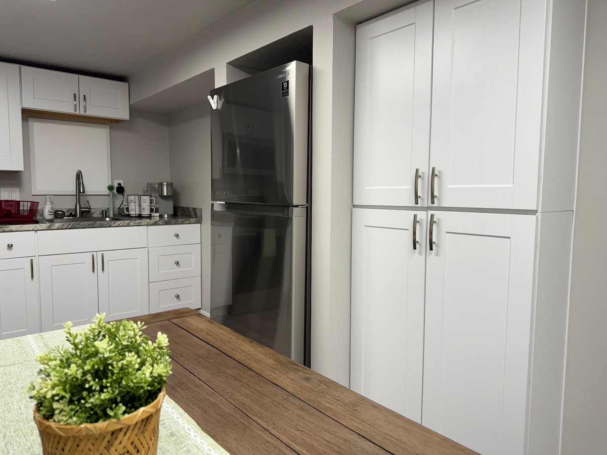 The kitchen features modern white cabinetry and a stainless steel refrigerator. A wooden dining table with a small potted plant sits in the foreground, while countertops are arranged with utensil holders and kitchen appliances.