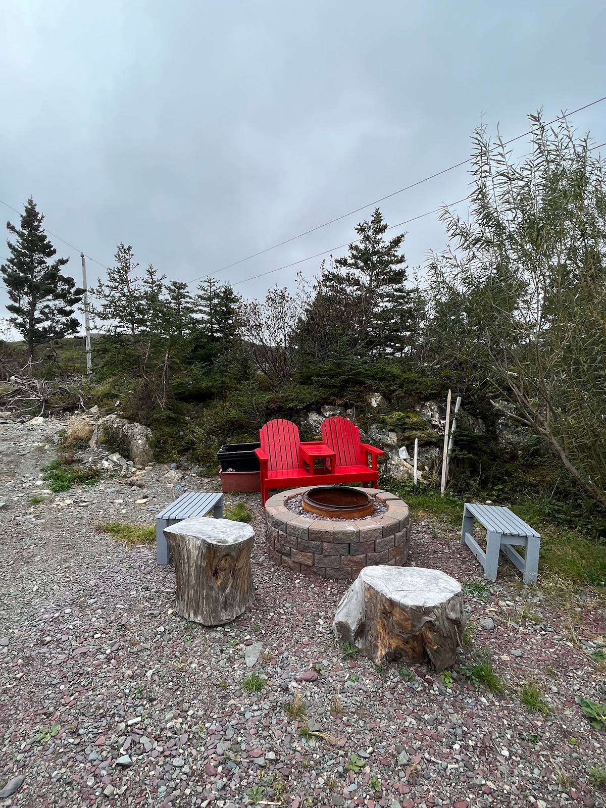 A designated outdoor area is displayed, featuring a circular stone fire pit surrounded by two red Adirondack chairs. Two rustic wooden stumps are positioned nearby along with two small benches, all set against a backdrop of natural greenery and rocky terrain.