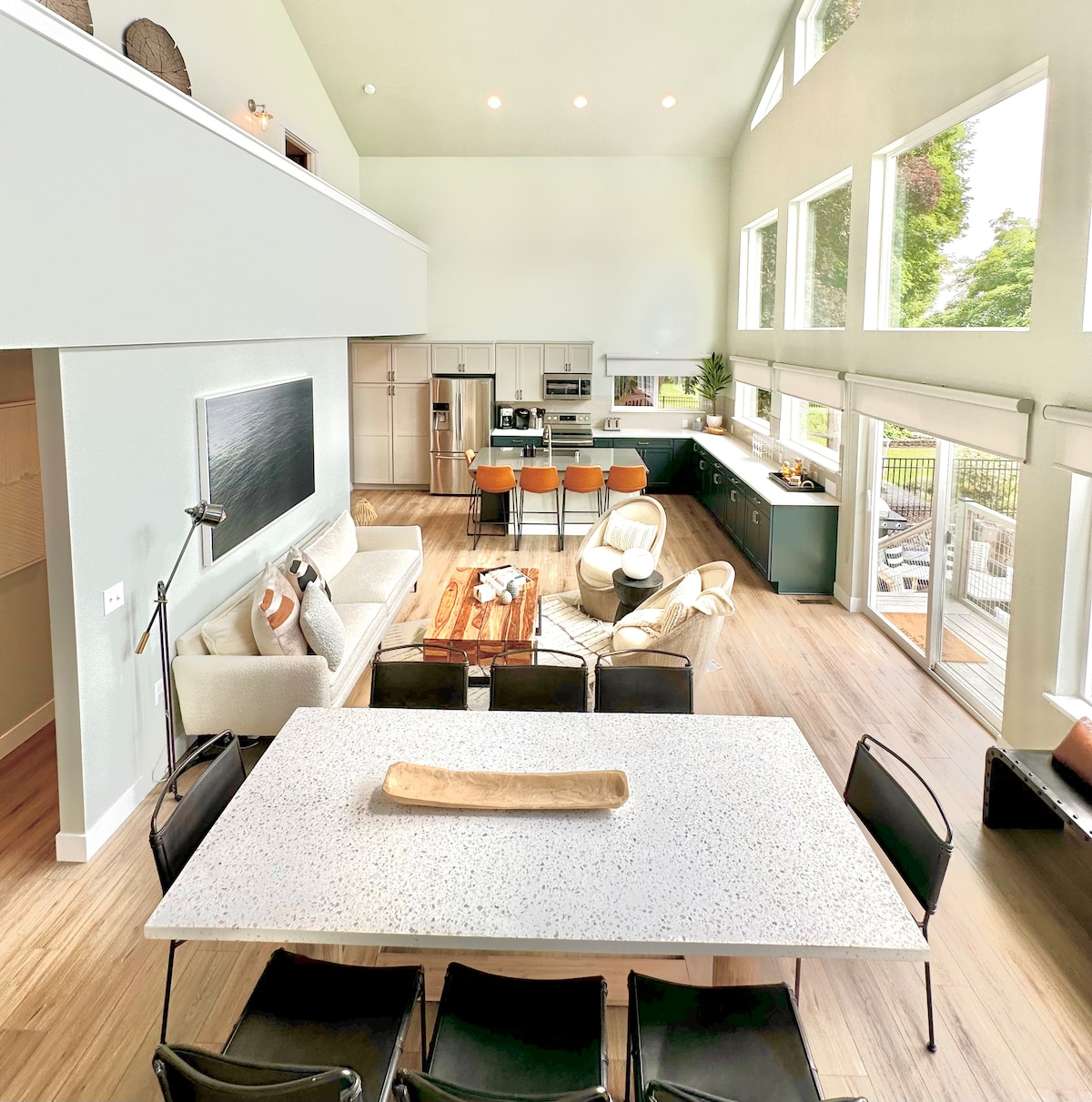 An open floor plan is showcased, featuring a dining area with a large speckled table and black chairs. The kitchen, equipped with modern appliances and bar seating, is visible in the background. Expansive windows allow natural light to fill the space, with greenery seen outside.