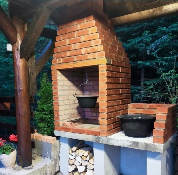 A charming outdoor cooking area features a sturdy brick barbecue with a chimney. A black pot hangs from a hook over the open flame, while a second pot rests on a flat surface nearby. Stacked firewood is neatly arranged beneath, surrounded by green foliage.