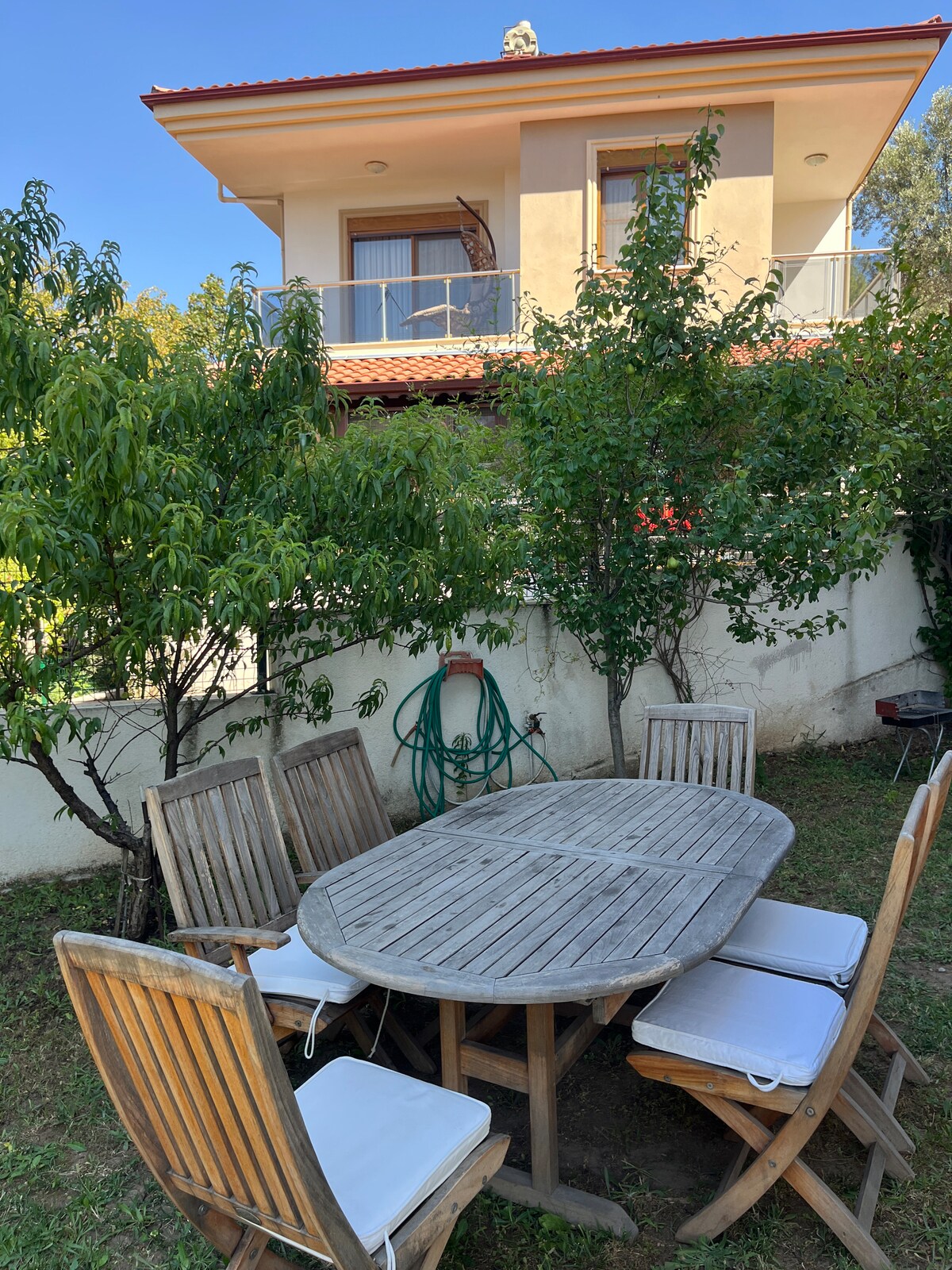 An outdoor dining area features a wooden oval table surrounded by six chairs, each with a cushion. Lush green trees provide dappled shade, while a house is visible in the background, showcasing a balcony with a simple railing.