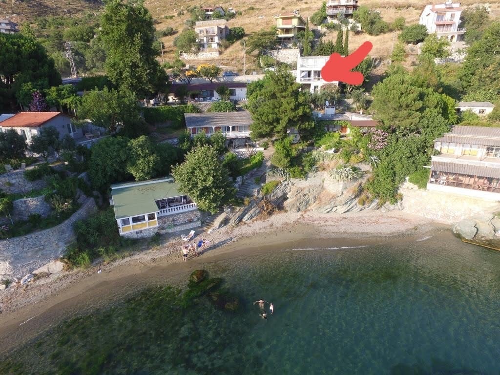 An aerial view captures the house surrounded by lush greenery and situated near a serene beach. The gentle waves lap at the sandy shore, while several buildings are visible along the coastline in the background, blending harmoniously with the landscape.