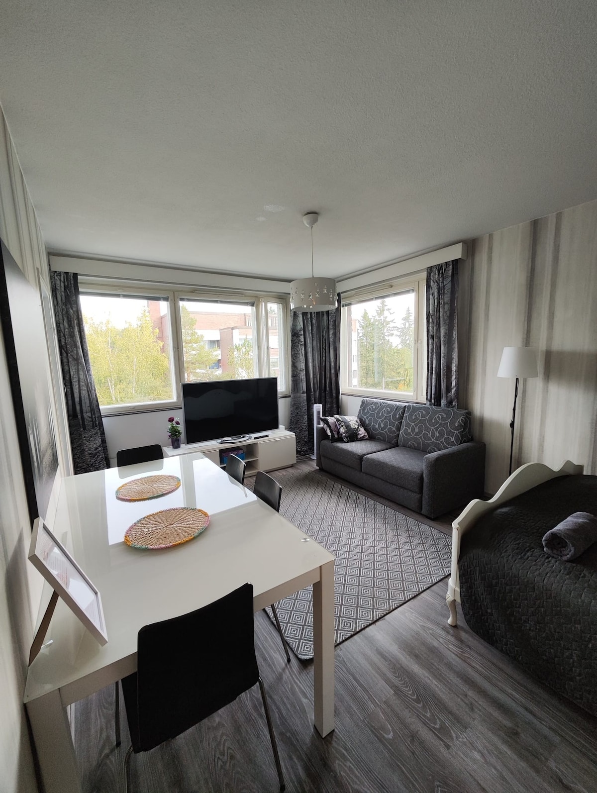 A compact living area encompasses a dining table set for two and a comfortable sofa arranged near large windows. Light streams in, highlighting a TV and decorative accents. A cozy bed features soft linens, and a subtle wall design adds depth to the space.