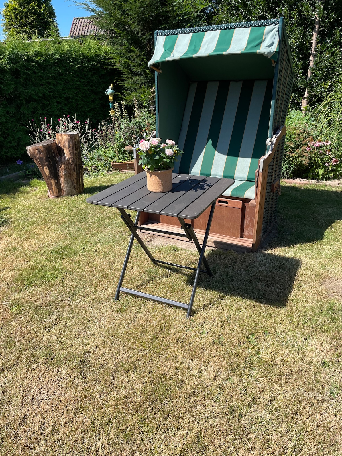 A beach chair featuring a striped green and white canopy is positioned in a grassy garden area. A small folding table with a potted flowering plant sits nearby, creating a relaxing outdoor space surrounded by greenery.