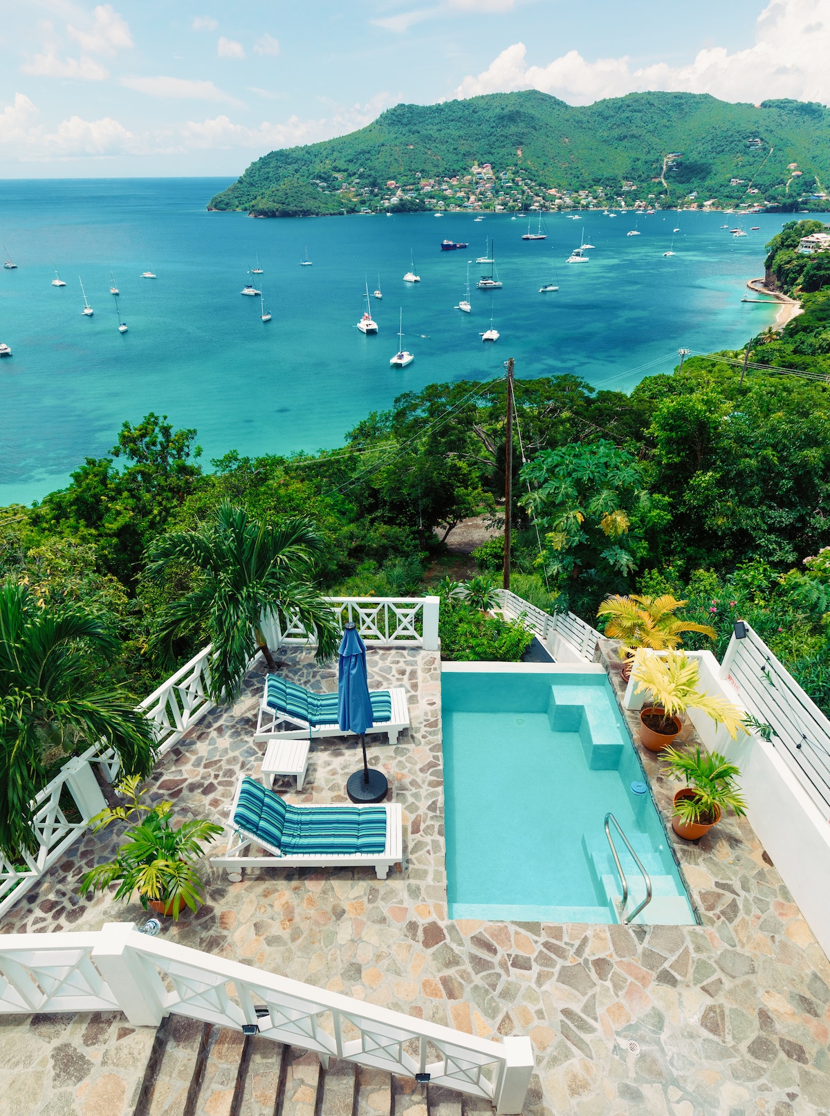 A serene outdoor area features a private pool surrounded by lush greenery. Lounge chairs with an umbrella offer comfort while enjoying the view of the bay and distant hillside, dotted with sailboats. Stone steps lead down from the terrace, enhancing the natural setting.