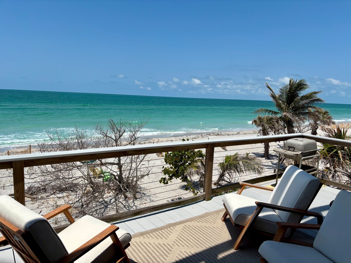 A spacious observation deck offers a relaxed seating area overlooking the beach. Two lounge chairs sit on a striped rug, while the ocean and palm trees are visible just beyond the railing, creating a serene coastal view under a clear blue sky.