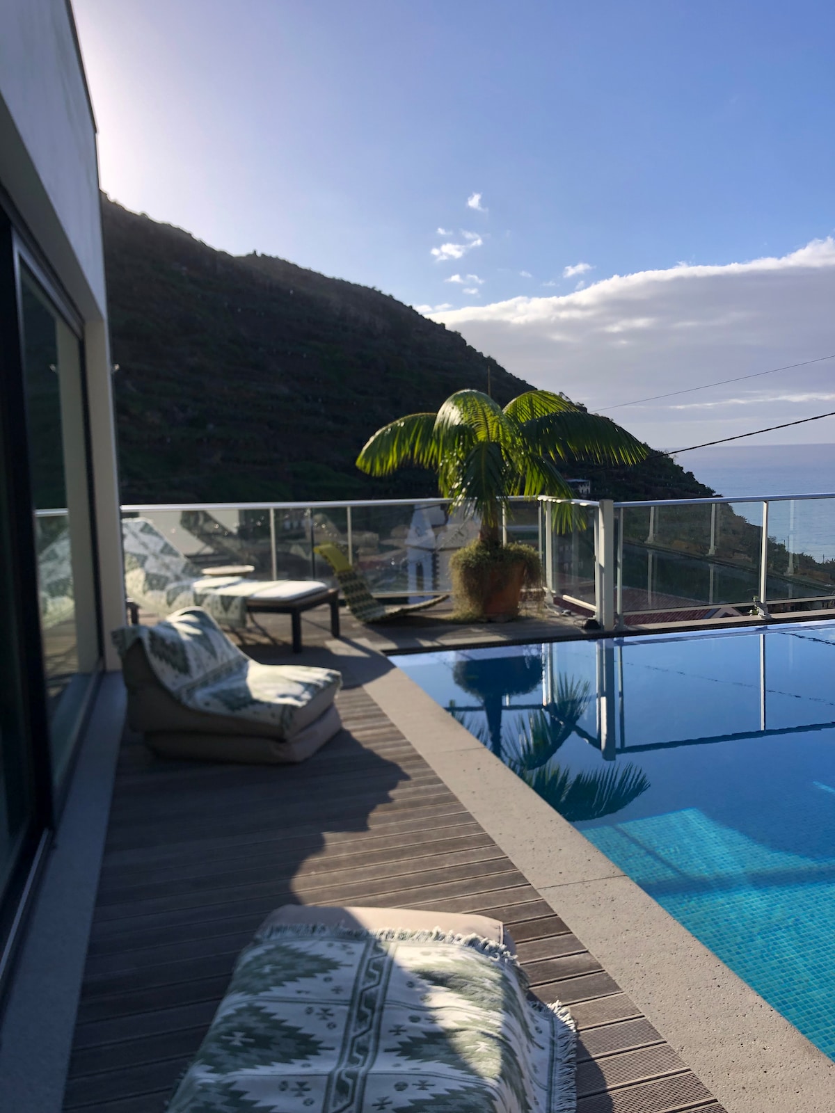 A private pool area is visible with a sunlit view of the surrounding hills. Comfortable outdoor seating is arranged nearby, complemented by a potted palm. The calm water reflects the clear sky, enhancing the serene atmosphere of the space.