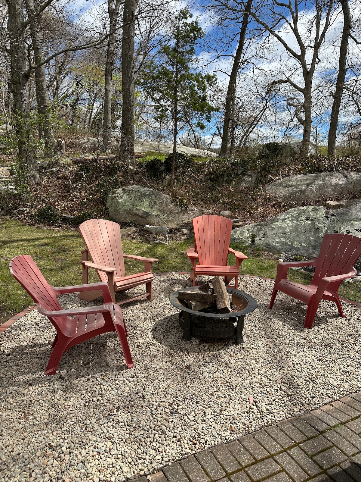 A fire pit is surrounded by four adirondack chairs, positioned on a gravel surface. The area is bordered by natural rock formations and trees, with a mix of sunlight and shade creating a serene outdoor setting.
