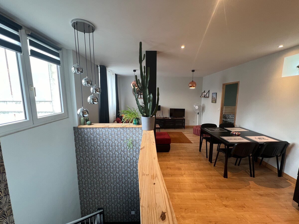 An open indoor space is viewed from above, showcasing a dining area with a black table and chairs adjacent to a cozy seating area featuring a red couch. Large windows allow natural light, while a tall cactus adds greenery to the space.