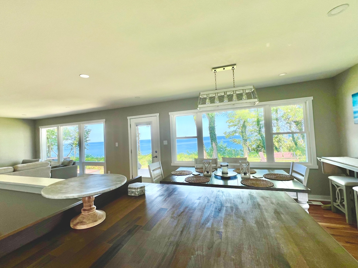 The dining area features a large wooden table surrounded by white chairs. Large windows provide a view of Lake Michigan and allow natural light to enter the space. The room's neutral walls complement the open layout leading to the living area.