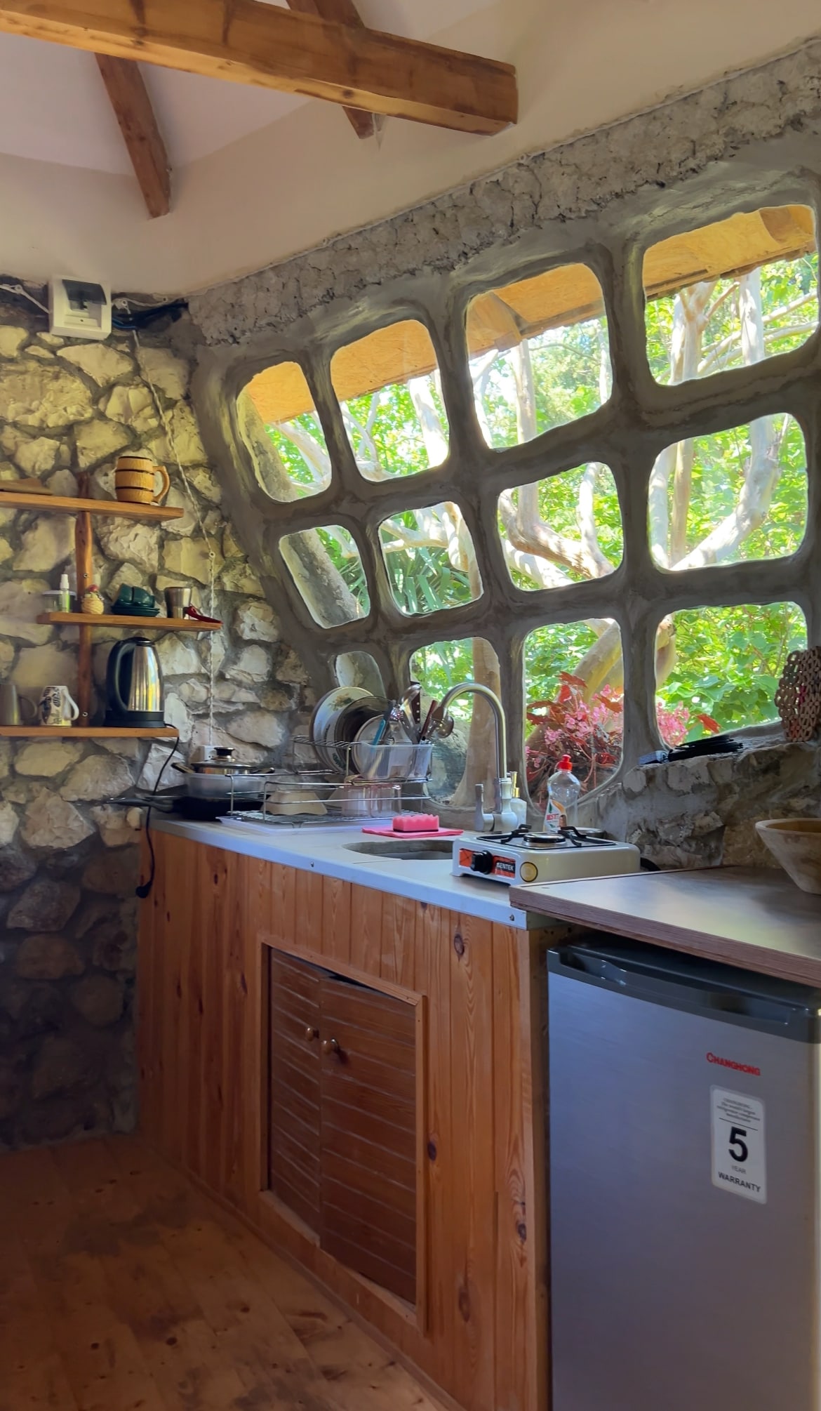 A rustic kitchen area features a stone wall and wooden cabinetry. A unique window design allows natural light to illuminate the space, surrounded by greenery. Kitchen appliances, including a stove and refrigerator, are arranged along the counter, creating a functional cooking environment.