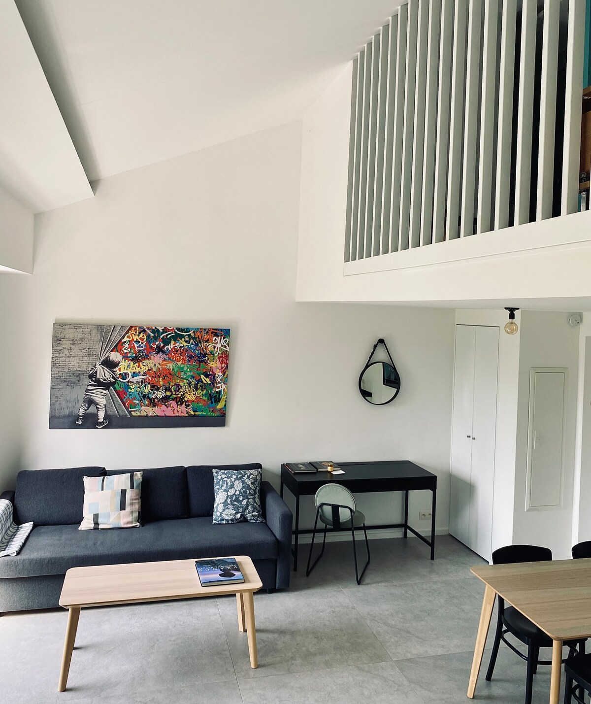 A spacious living area is presented with a modern design, featuring a dark sofa and a light wooden coffee table. A colorful wall art piece decorates the space, while a mirror hangs near a door. A dining area with chairs is positioned in the corner.