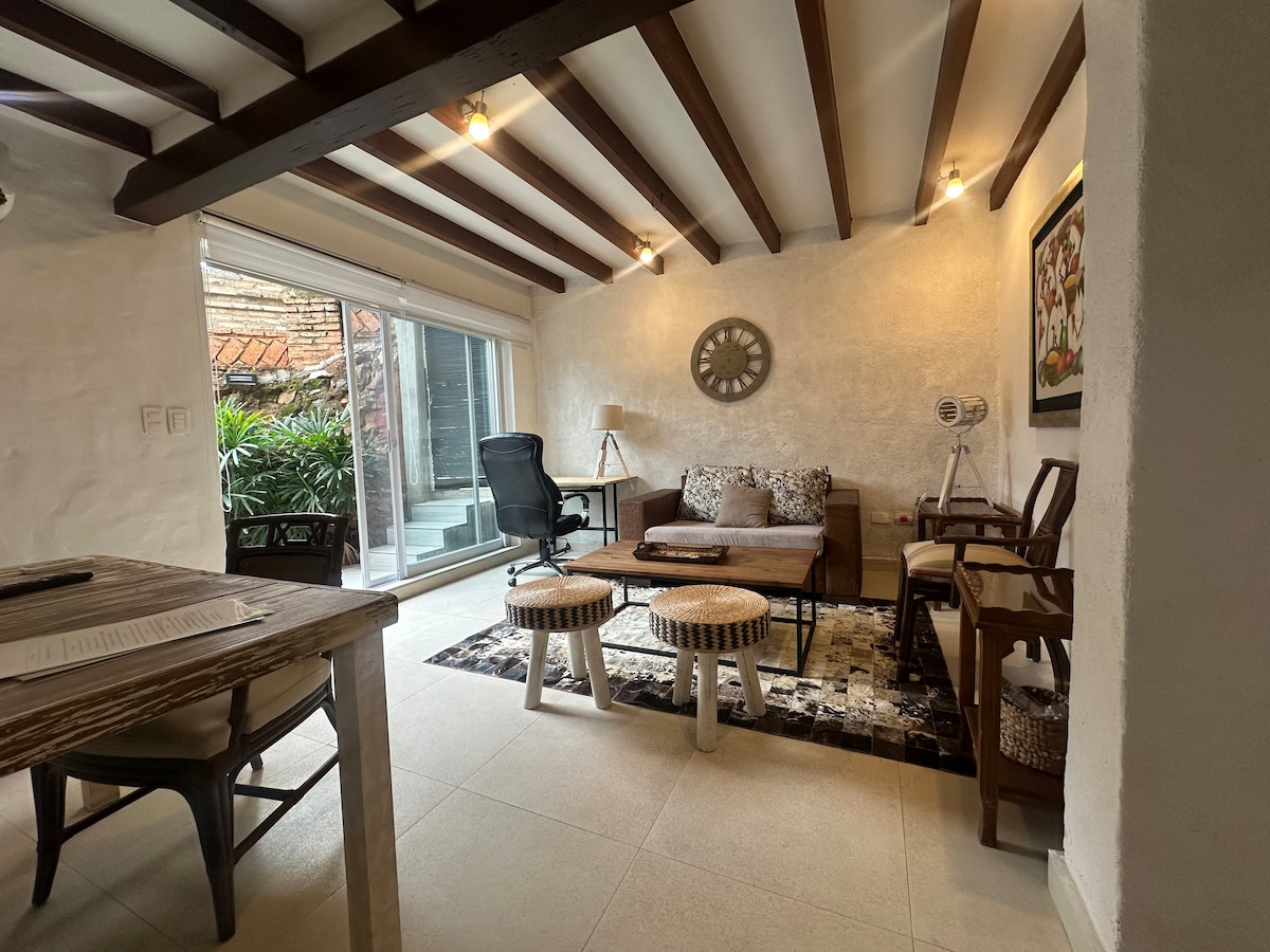 The living area features a mix of comfortable furnishings, including a sofa, two accent chairs, and a coffee table. Large glass doors provide natural light and a view of the private patio with greenery, enhancing the space's brightness. A wall clock and artwork add to the decor.