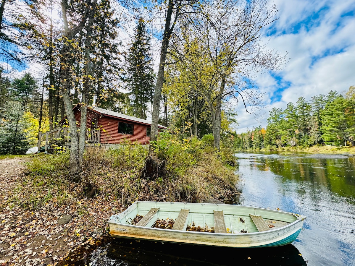 A riverside cabin with a deep red exterior is framed by trees and autumn foliage. A small rowboat rests on the water's edge, surrounded by fallen leaves. The calm river reflects the surrounding landscape and clouds, enhancing the natural beauty of the setting.