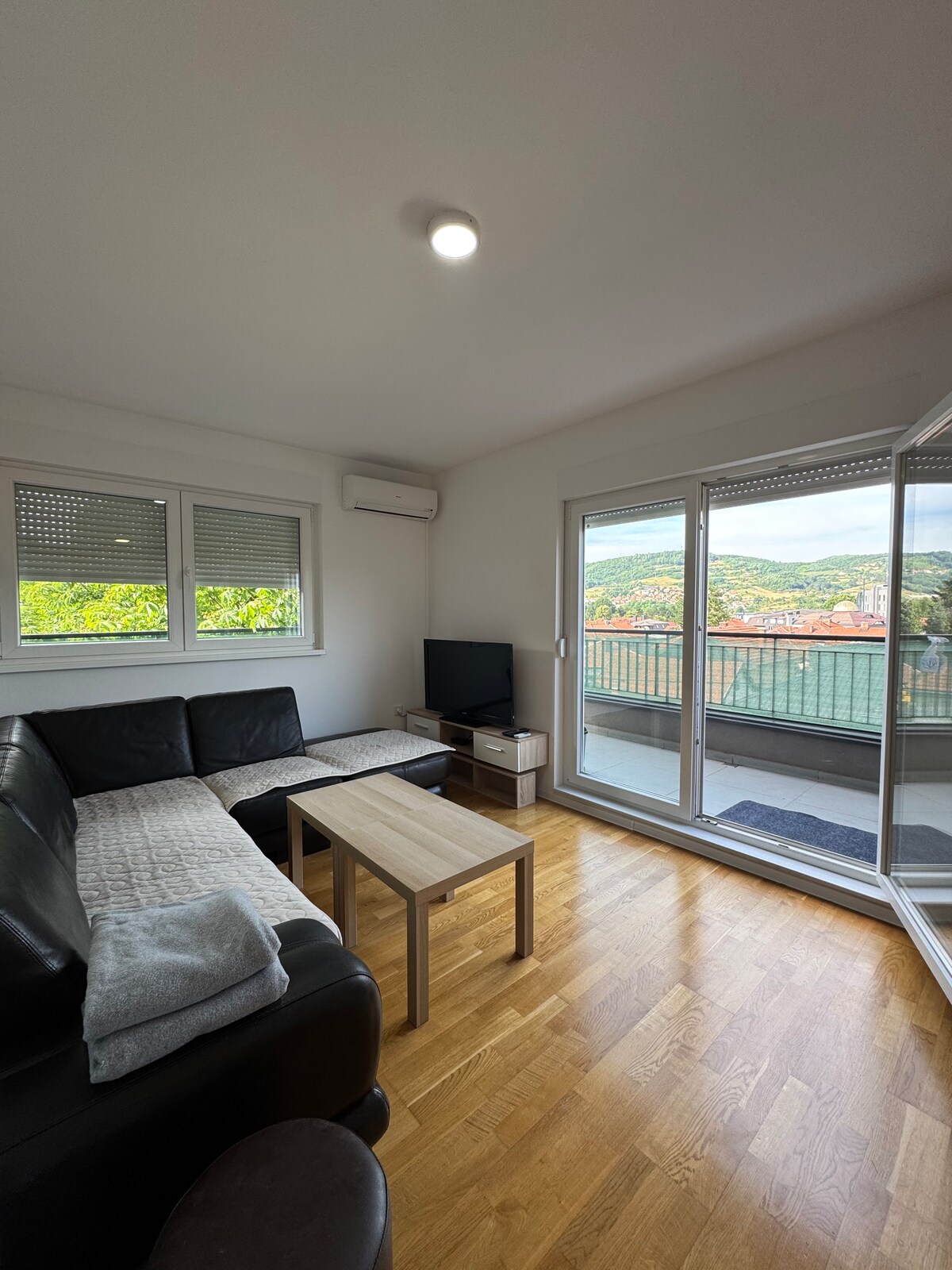 A bright living area features a comfortable black couch with light-colored throws and a wooden coffee table at its center. Large glass doors open to a balcony, offering views of the surrounding landscape. A television is positioned near the window, enhancing entertainment options.