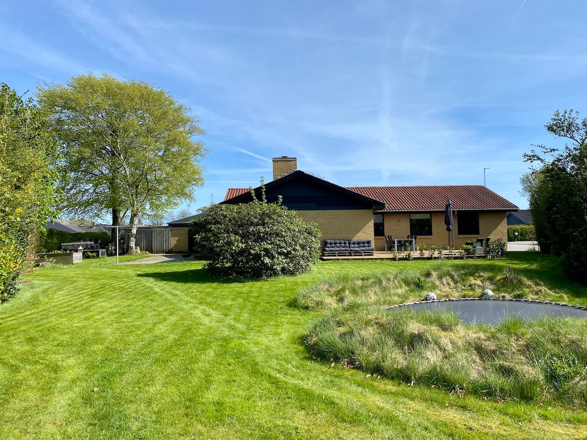 A single-story house is set in a green yard, surrounded by well-maintained grass and shrubs. A small pond is visible in the foreground, alongside a seating area. The structure features a combination of large windows and a red-tiled roof, contributing to a serene outdoor environment.