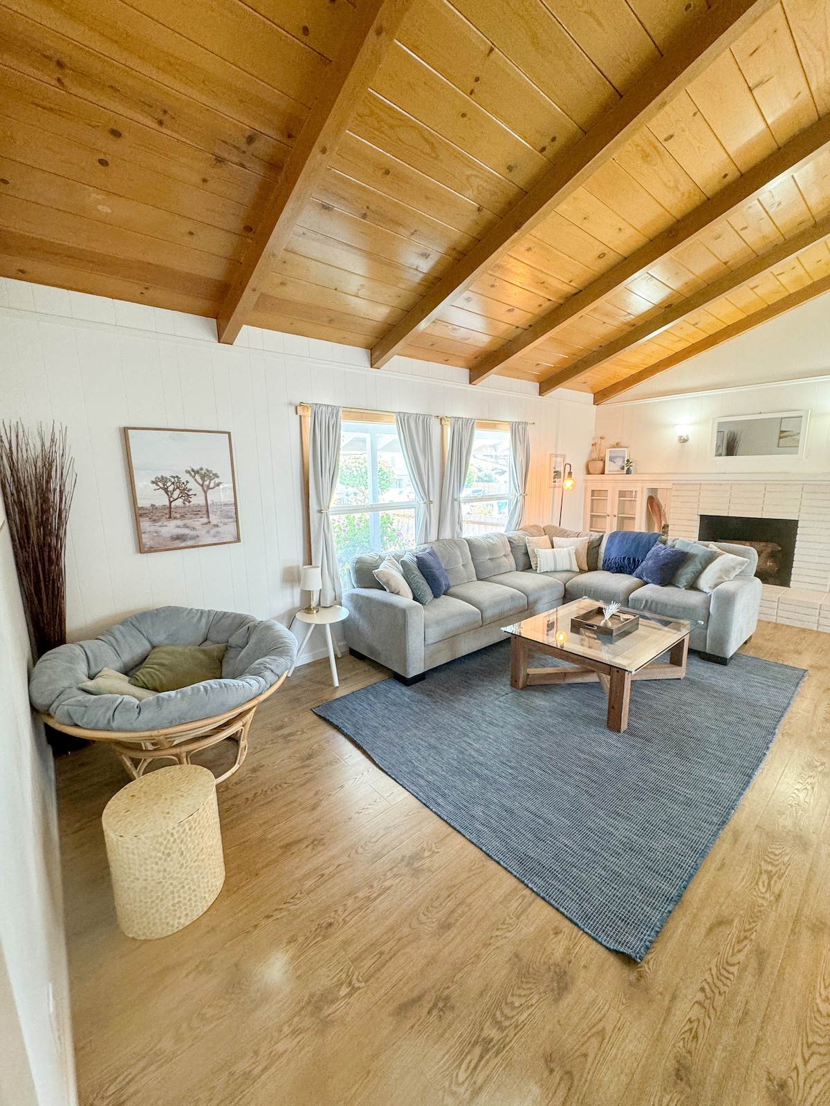 A bright living room features a vaulted wooden ceiling and large windows. A comfortable gray sofa is arranged around a central coffee table on a textured area rug. A cozy chair and a small decorative table add warmth, with a wall art piece enhancing the inviting space.
