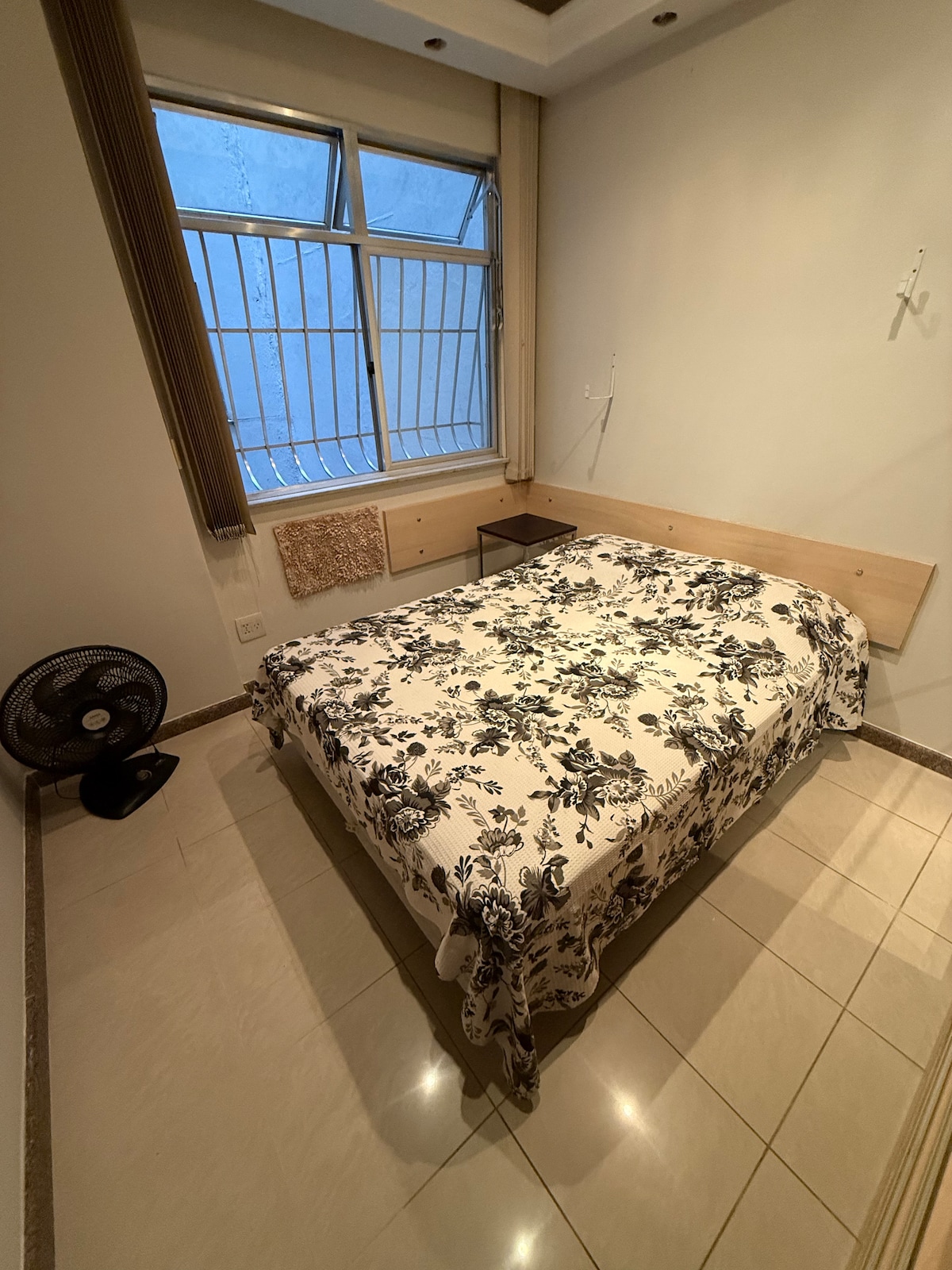 A bedroom is featured with a spacious bed covered in a floral patterned blanket. Light streams in through a large window with a grid design, illuminating the tiled floor and minimalist furnishings, including a small table and a fan positioned nearby.