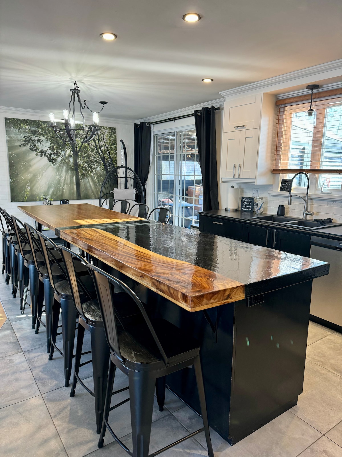 A modern kitchen space features a large, glossy wooden countertop that serves as a bar with multiple high black chairs. Natural light filters through large windows covered with dark curtains, and a nature-themed wall art adds a serene touch. Sleek cabinets and fixtures complement the design.