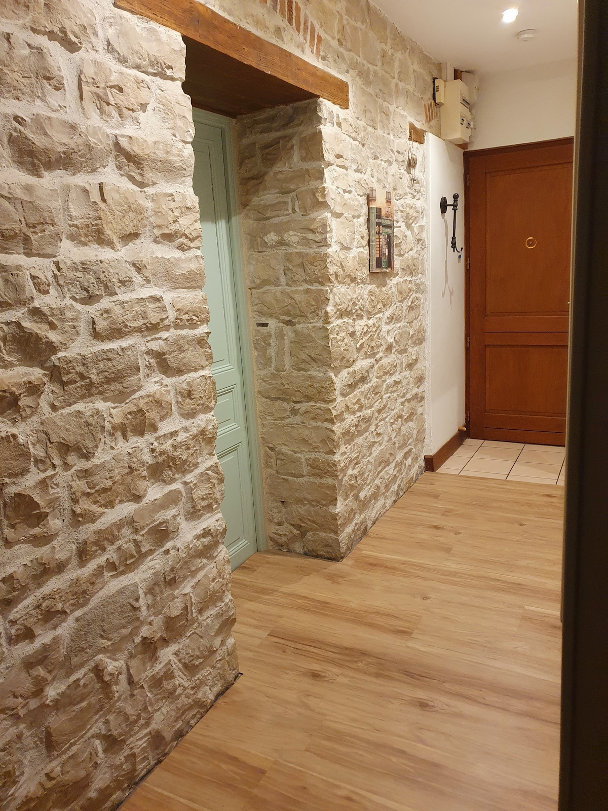 An inviting hallway is framed by textured stone walls, showcasing a blend of rustic charm and modern design. A light wooden floor extends throughout, while a green doorway and a wooden entry door provide access to various rooms. Soft lighting enhances the warm atmosphere.