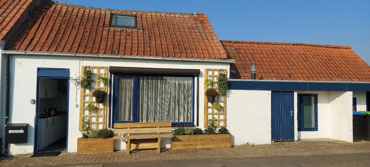 The exterior of the charming dike house features a red-tiled roof and white walls. A wooden bench is placed in front of a large window adorned with planters. The entrance is accented by a blue door, while the clear sky enhances the serene ambiance.