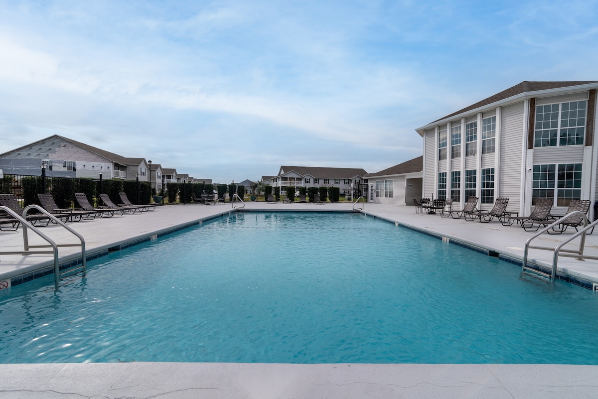 A spacious outdoor pool is central to the large courtyard, surrounded by lounge chairs and greenery. The pool's clear blue water reflects the sky, while modern buildings provide a contemporary backdrop, enhancing the inviting atmosphere of the communal area.