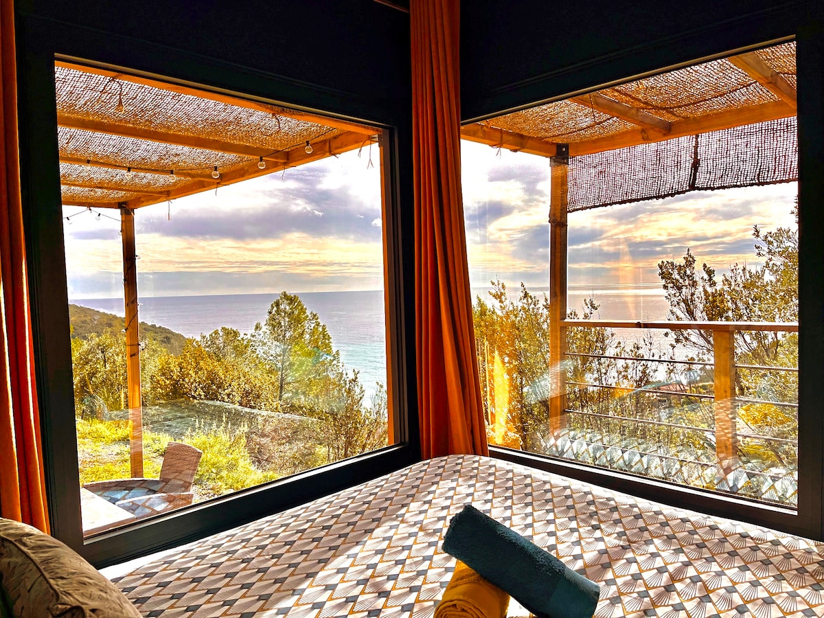 Large windows frame a serene view of the sea and surrounding greenery. Soft orange drapes complement the natural light, while a neatly arranged bed features a patterned duvet and a rolled towel. A peaceful outdoor terrace is visible just beyond the glass.