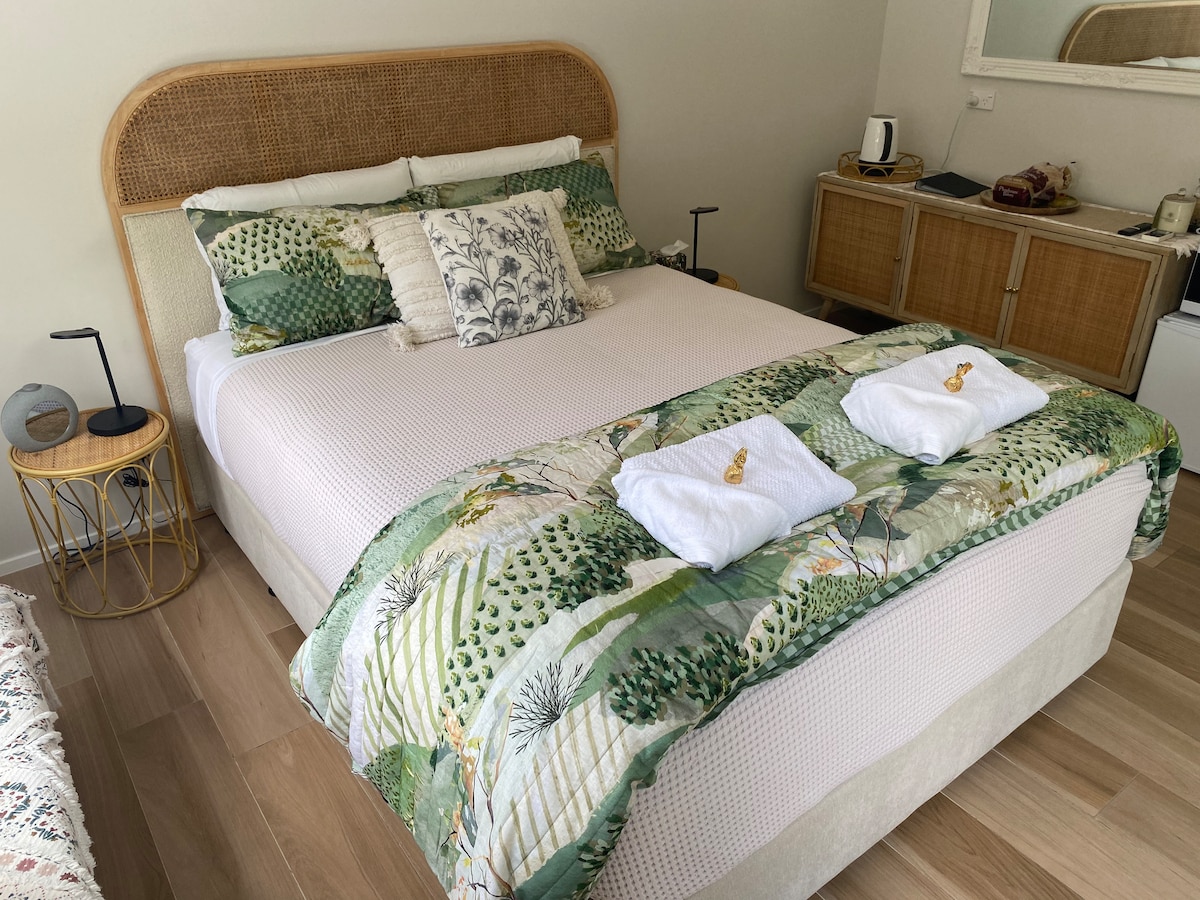 A spacious bed is dressed in a green and white patterned duvet, accompanied by neatly arranged white towels at the foot. Decorative pillows add texture, while a bedside table holds a small lamp. A dresser and a kettle are visible in the background.