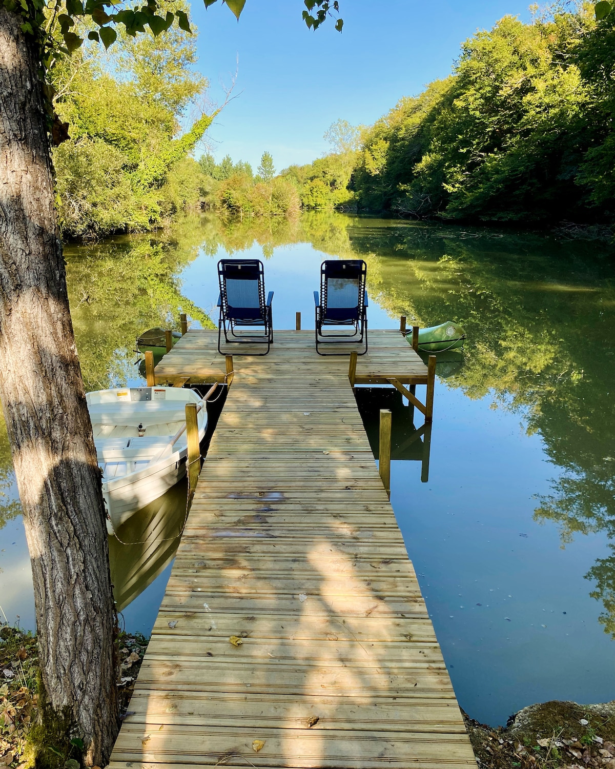 A wooden dock extends over the calm waters of the lake, flanked by vibrant greenery. Two relaxing chairs are positioned for tranquil views, and a small rowboat is tied nearby. The serene landscape is reflected in the water, enhancing the natural ambiance.