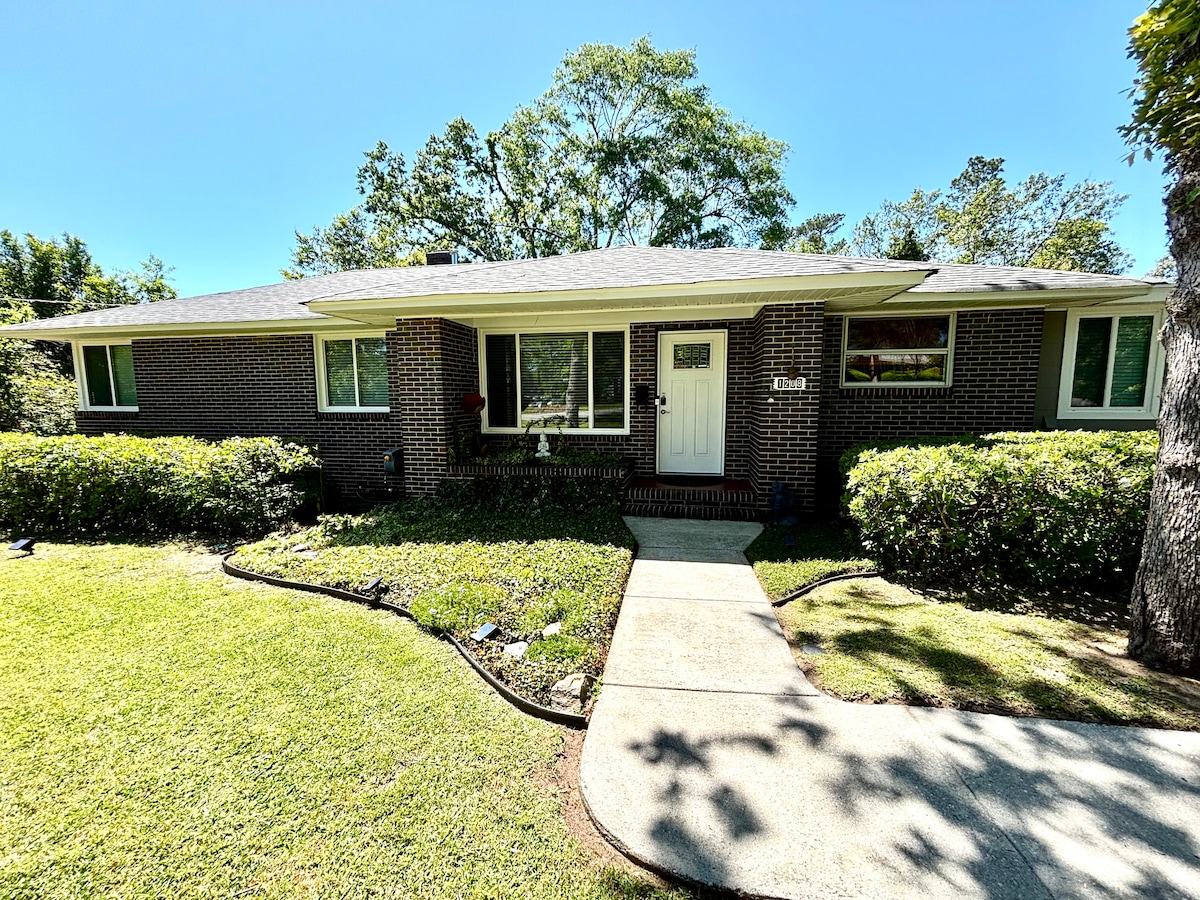 A mid-century house is showcased with a brick facade framed by well-maintained greenery. A pathway leads to the entrance, bordered by manicured lawns. Large windows allow natural light to illuminate the entryway, enhancing the welcoming appearance of the home.