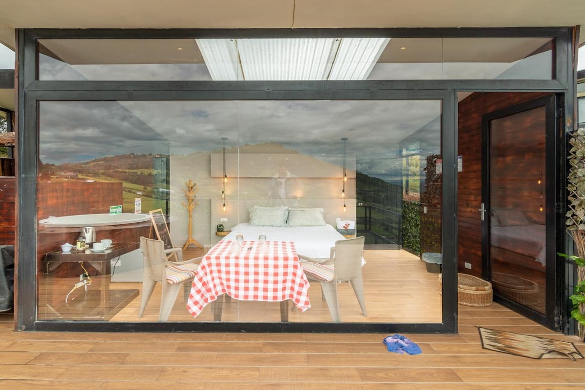 A cozy room showcases a spacious bed, elegantly framed by large glass walls. A dining table set with a checkered tablecloth is positioned near the entry, while a small kitchenette is visible to the side. Natural light brightens the interior, enhancing the inviting atmosphere.