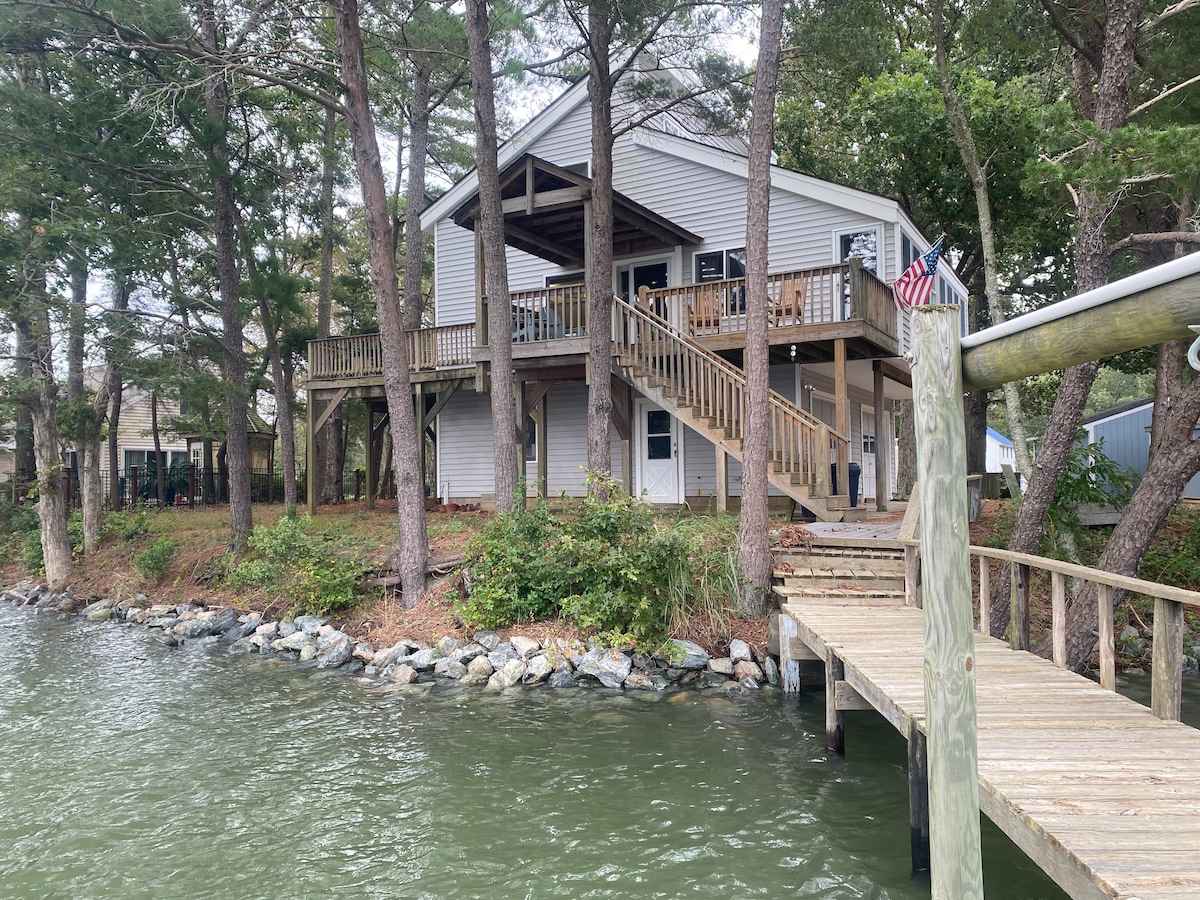 The exterior of a two-story house is displayed, featuring a wraparound deck and steps leading to a private dock. Surrounding trees provide shade, while the water is visible in the foreground. An American flag is seen on the deck railing.