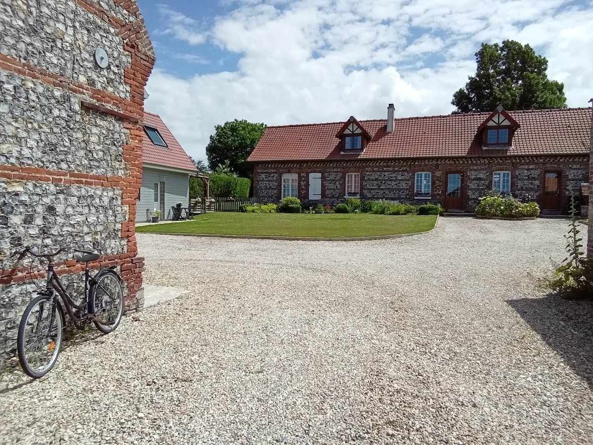 The courtyard of an old farmhouse is visible, showcasing a gravel surface and green grass. Stone and brick buildings line the area, with a few bicycles parked nearby. A clear sky and scattered clouds create a serene backdrop.