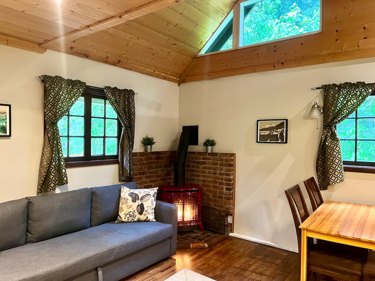 A cozy living area features a gray couch adorned with a decorative pillow. A warm wood stove is situated against a brick wall, providing a comforting focal point. Natural light enters through large windows, framed by patterned curtains that complement the wooden ceiling.