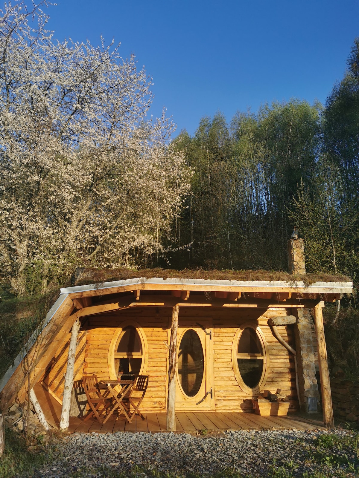 A uniquely crafted wooden cottage is surrounded by nature, featuring a sloped roof and rounded front door. A small terrace with two wooden chairs and a table is present, inviting guests to relax. Blossoming trees add a touch of greenery to the scenic landscape.