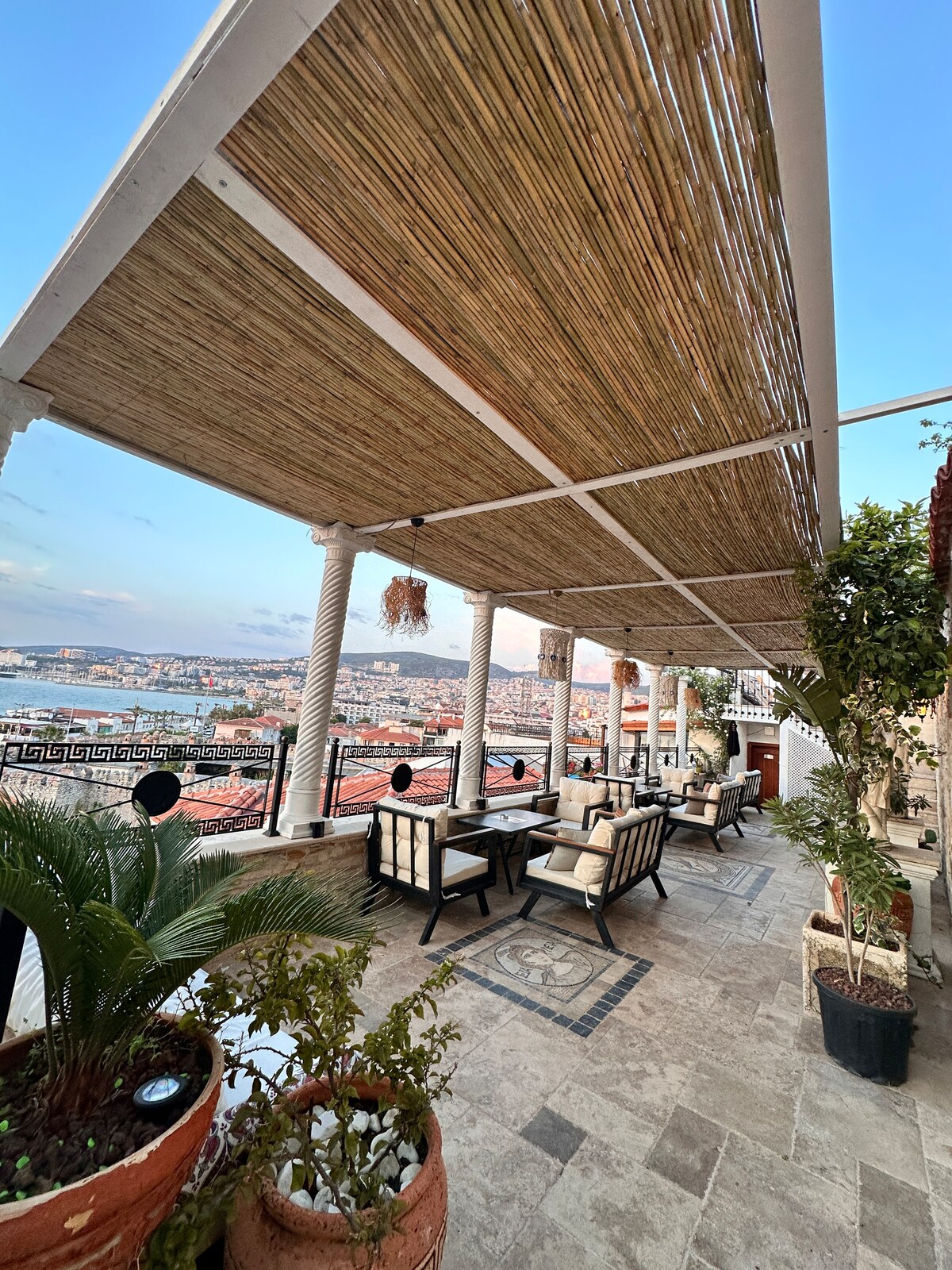 A spacious terrace is adorned with comfortable seating arrangements, surrounded by plants in terracotta pots. The area is covered with a striped canopy, providing shade. Scenic views of the surrounding cityscape and coastline can be appreciated from this relaxing outdoor space.