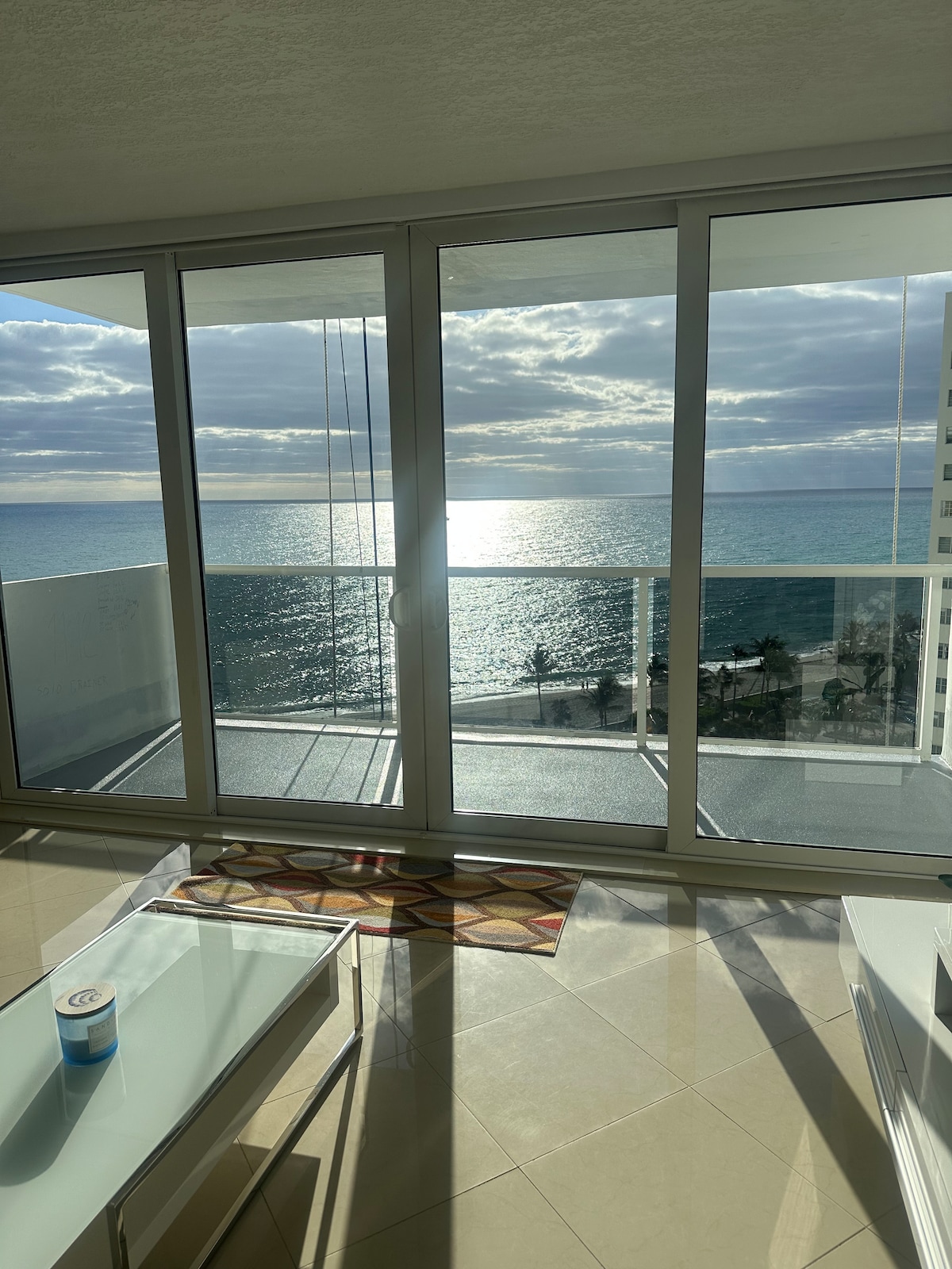 A large glass sliding door opens up to a breathtaking view of the ocean, with sunlight reflecting on the water's surface. The space is filled with natural light, showcasing a modern, open layout. A colorful area rug adds a touch of comfort to the room.