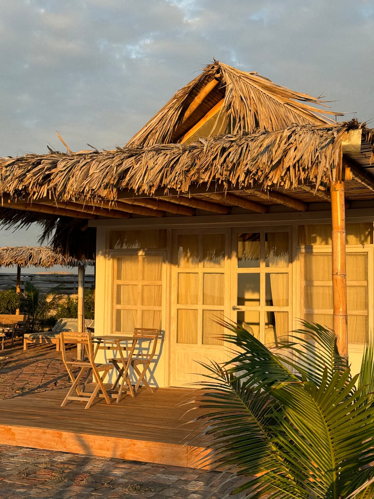 A charming beach cabin showcases a thatched roof and large wooden windows. A small wooden deck features a bistro table and two chairs, surrounded by tropical plants. The warm light of the setting sun enhances the cabin's inviting exterior.