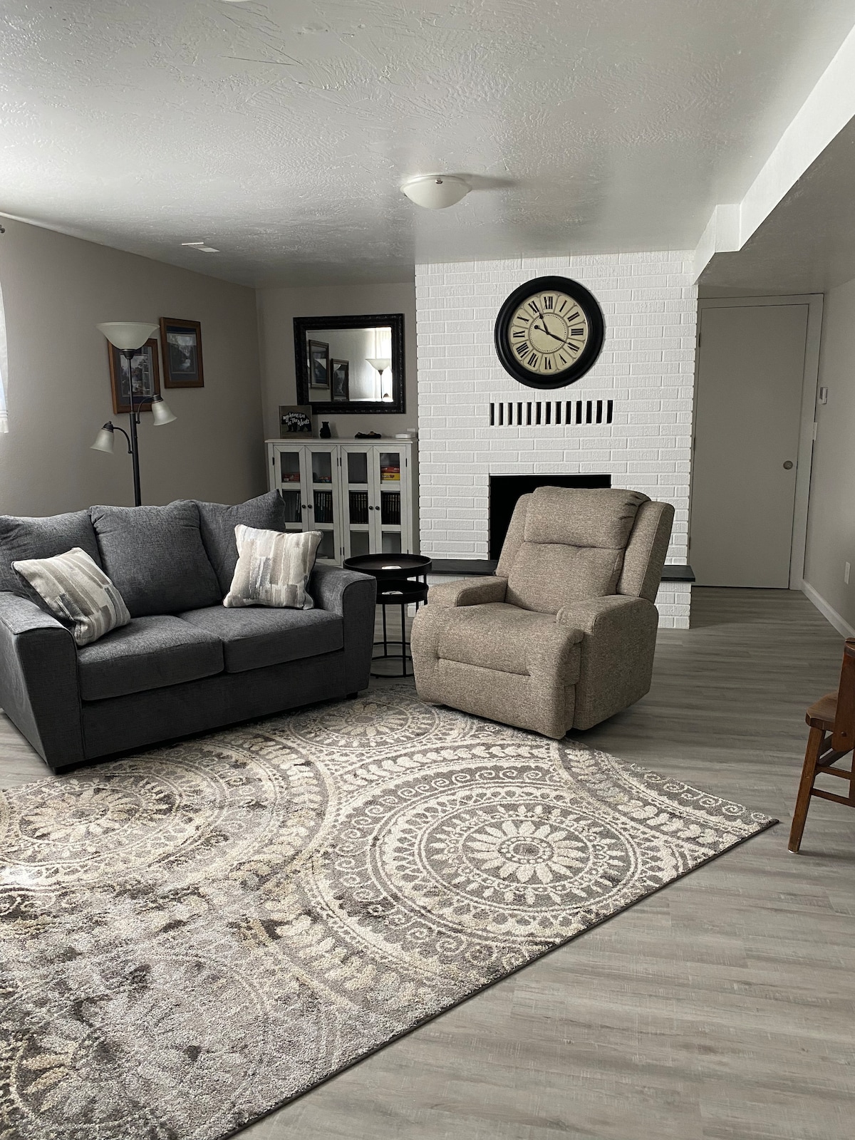 The living room is furnished with a gray sofa and a recliner, positioned on a patterned area rug. A white brick fireplace is visible in the background, complemented by a large wall clock. Natural light filters through nearby windows.