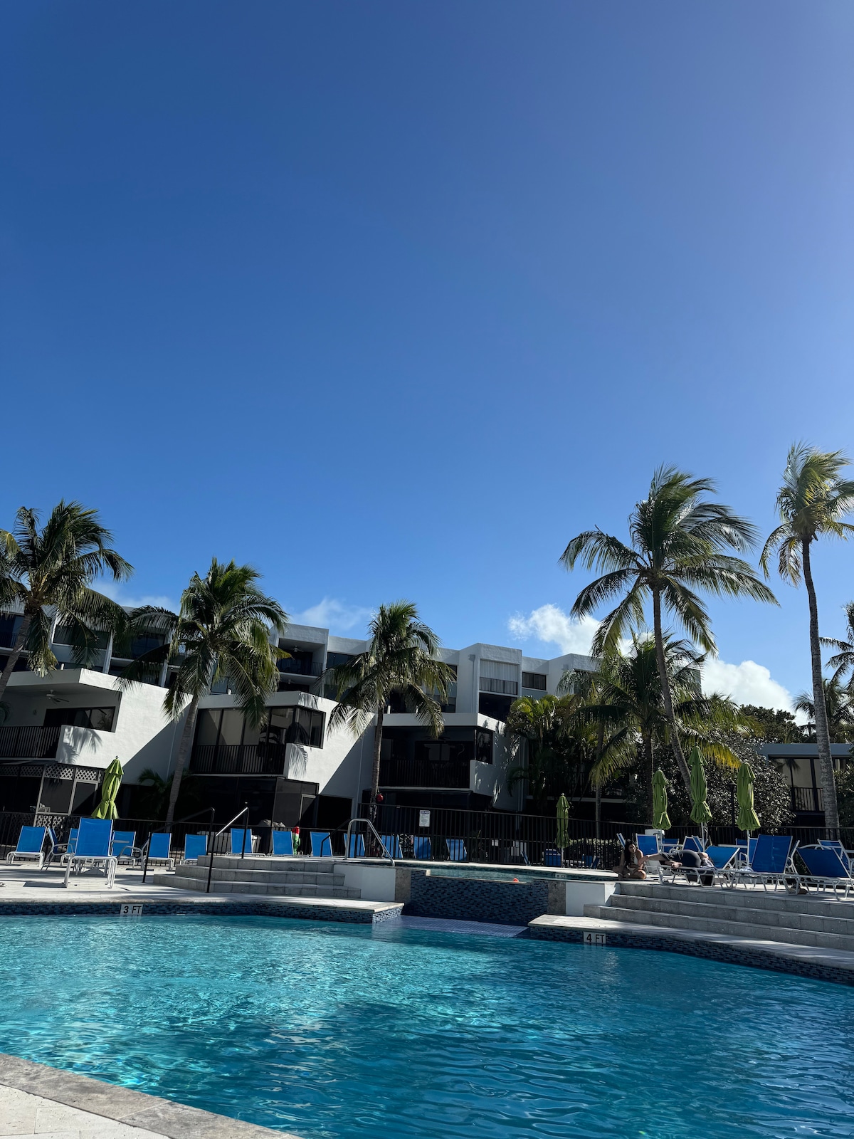 A clear blue sky hovers over a swimming pool, surrounded by lounge chairs and vibrant palm trees. The two-story buildings of the complex provide a backdrop, with a mix of greenery and seating areas visible around the pool's edge.
