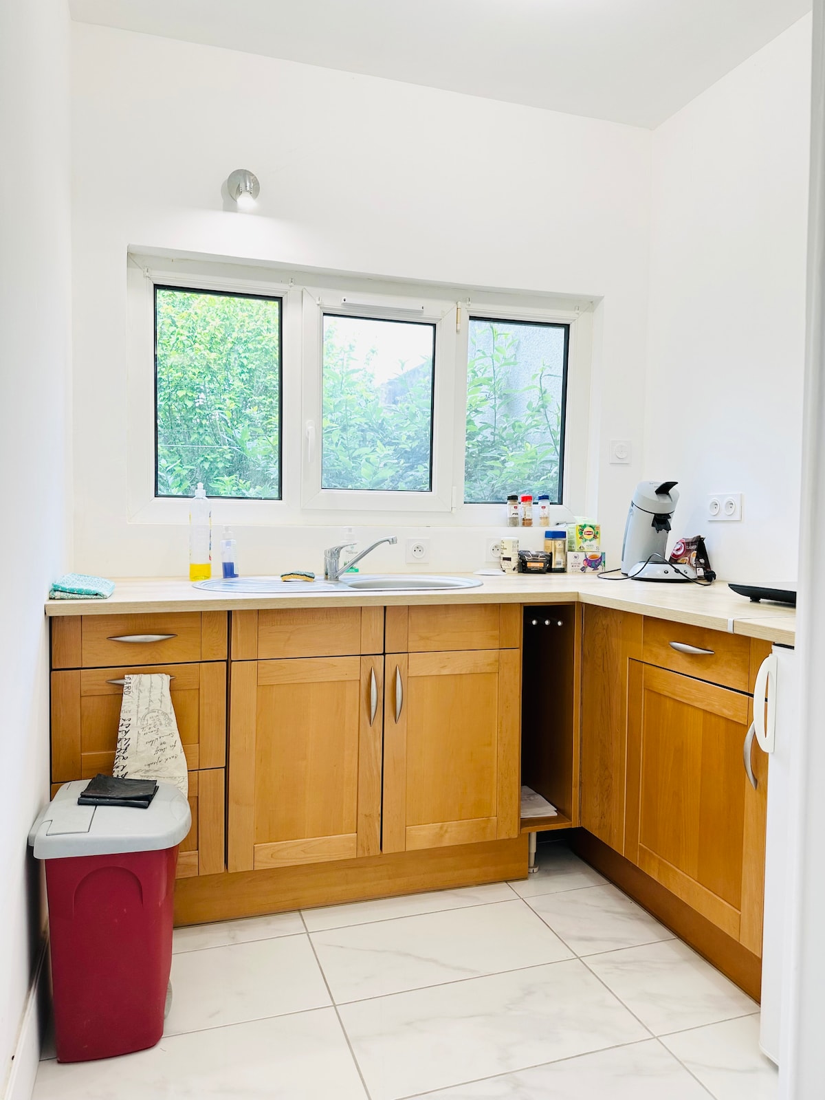 A modern kitchen with wooden cabinetry and a clean countertop is illuminated by natural light streaming through a window. The sink area features a small appliance and various condiments on display. Light-toned tiles create a bright and open atmosphere.