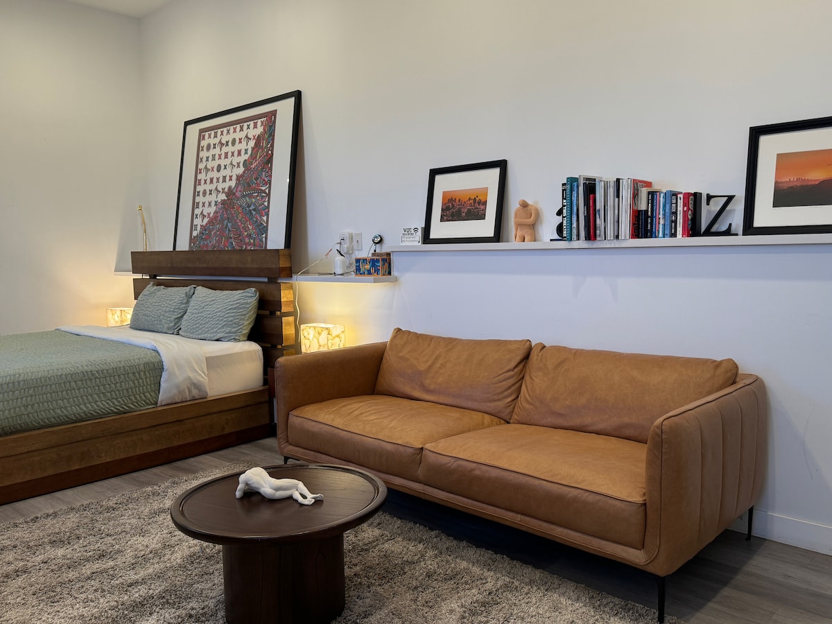 A spacious living area features a comfortable brown sofa with a circular table in front. Behind, a bed is positioned against the wall, adorned with light blue bedding. Shelves display a collection of books and framed photographs, complemented by soft lighting from wall fixtures.