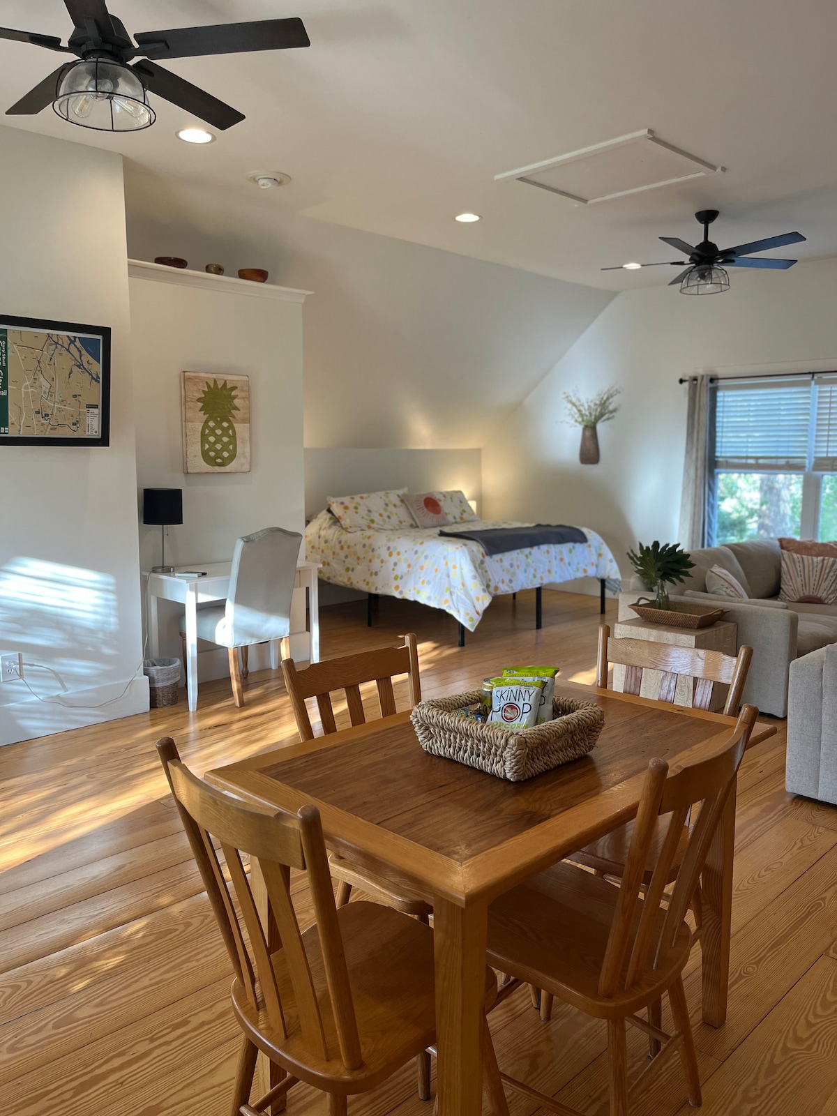 A bright, open living space features wide plank flooring, a king bed draped with a yellow floral patterned blanket, and two comfortable seating areas. A wooden dining table sits in the center, surrounded by four chairs. Natural light filters through the window drapes, creating a warm ambiance.