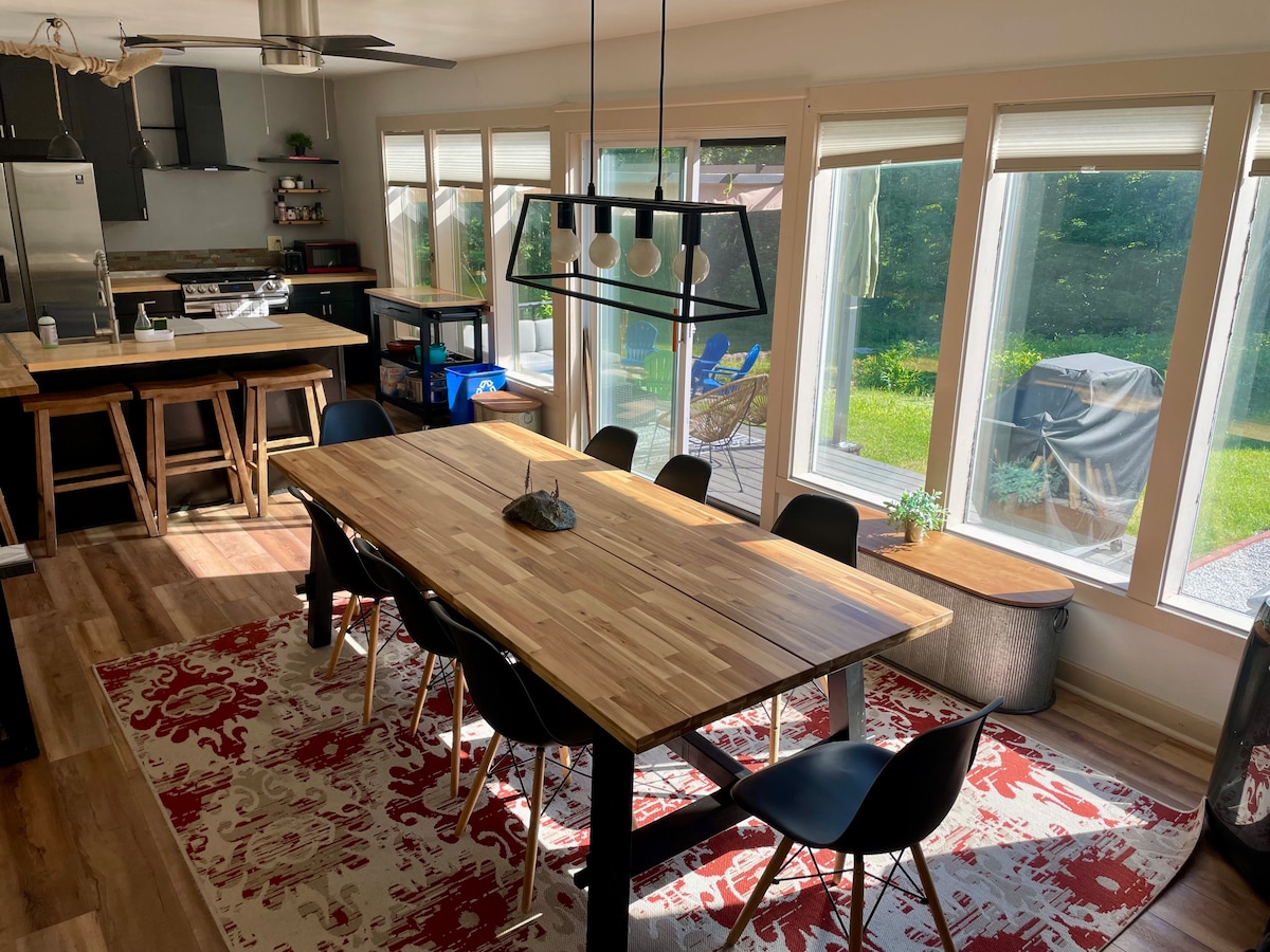 A bright dining area features a large wooden table surrounded by six black chairs. Sunlight filters through large windows, illuminating the space. A patterned rug adds warmth to the wood flooring, while a sleek light fixture hangs above the table, enhancing the inviting atmosphere.
