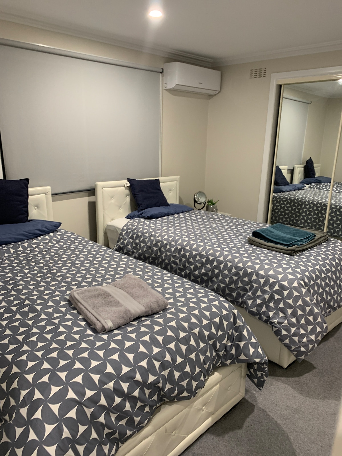A well-lit bedroom features two spacious beds dressed in patterned bedding. Each bed is complemented by navy blue pillows. A mirror reflects the room's layout, while a small plant and clock add subtle decor on the bedside table. Natural light is filtered by a window shade.