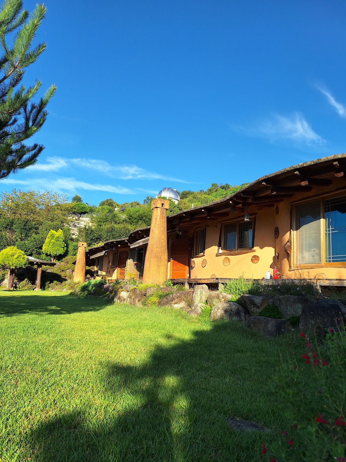 A rustic structure with natural wood accents and tall stone chimneys is featured under a clear blue sky. Lush green grass surrounds the building, with trees and shrubs in the background, creating a peaceful outdoor setting.