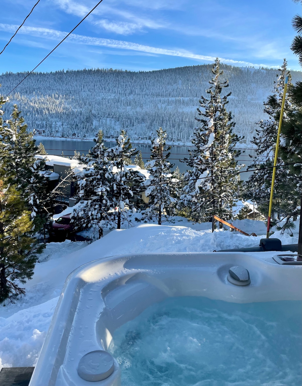 A hot tub is visible, surrounded by snow-covered trees and a view of distant mountains under a clear blue sky. The steaming water invites relaxation as winter scenery enhances the tranquil atmosphere.